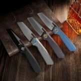 Gruppenaufnahme der vier Vosteed Xeno Varianten (Modell A3801) mit Titangriffen und Vanax-Klingen in verschiedenen Finishes: Black Ti-PVD, Dim Gray Acid Wash, Dark Reddish Purple und Blue.